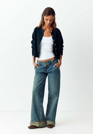 Frau trägt einen schwarzen, geknöpften Cardigan über einem weißen Tanktop, weit geschnittene, aufgekrempelte Jeans und braune Schuhe mit Punkten, steht mit Händen in den Taschen.