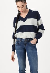 Jersey de punto a rayas en azul marino y gris con escote en V, combinado con jeans de mezclilla azules. El jersey cuenta con mangas largas y puños acanalados.