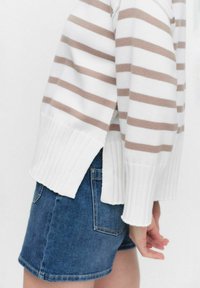 Gestreepte gebreide trui in wit en beige, met een geribde onderkant en zijsplitten, gecombineerd met blauwe denim shorts. Zachte textuur, casual ontwerp.