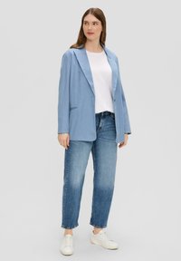 Blazer bleu clair avec revers crantés, un bouton et deux poches, associé à un t-shirt blanc et un jean bleu, porté avec des baskets blanches.