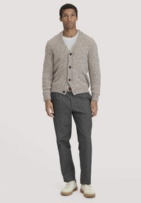 Lichtbruine gebreide cardigan met een V-hals, met donkere knopen, gedragen over een wit t-shirt, gecombineerd met grijze broek en beige sneakers.