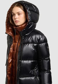 Svart vadderad jacka med blank yta, oversized huva och kontrasterande brunt foder. Har dragkedja och justerbara dragsnören.