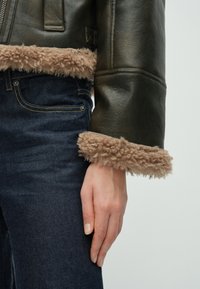 Giacca in pelle verde scuro con rifiniture di pelliccia beige soffice ai polsini, abbinata a jeans di denim scuro. Primo piano di una mano appoggiata sui jeans.