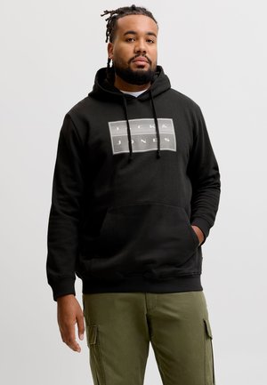 Mężczyzna z zaplecionymi włosami i brodą, ubrany w czarną bluzę z kapturem Jack & Jones i zielone spodnie typu cargo, stojący na tle jednolitego jasnego tła.