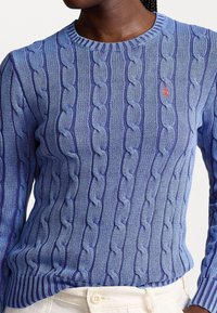 Blå kabelstickad tröja med rund halsringning. Har varierande nyanser av blått och en liten röd logotyp på bröstet. Texturerat mönster över hela tröjan.