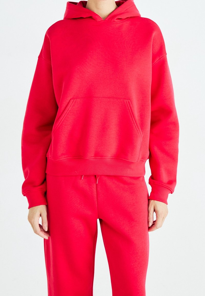 Personne portant un sweat à capuche rouge vif avec une poche avant et un pantalon de survêtement rouge assorti, debout devant un fond blanc.