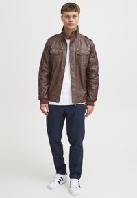 Veste en cuir marron avec deux poches poitrine, poignets côtelés et col montant, associée à un pantalon navy et des baskets blanches.