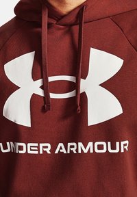 Bordowy hoodie z dużym białym logo Under Armour i tekstem. Wykonany z miękkiego materiału o teksturowanym wykończeniu, zawiera regulowane sznurki.