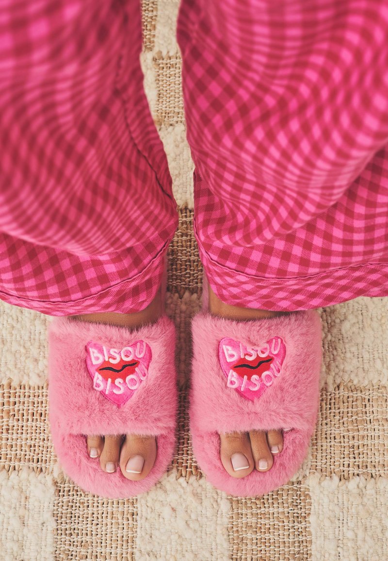 Chaussons en peluche roses avec un patch en forme de cœur comportant le texte "BISOU" et un graphisme de rouge à lèvres. Portés avec un pantalon de pyjama à carreaux roses.