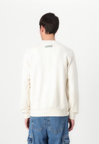 Cream sweatshirt with a round neck and long sleeves, featuring a small green "Lacoste" label on the back, paired with blue denim shorts.