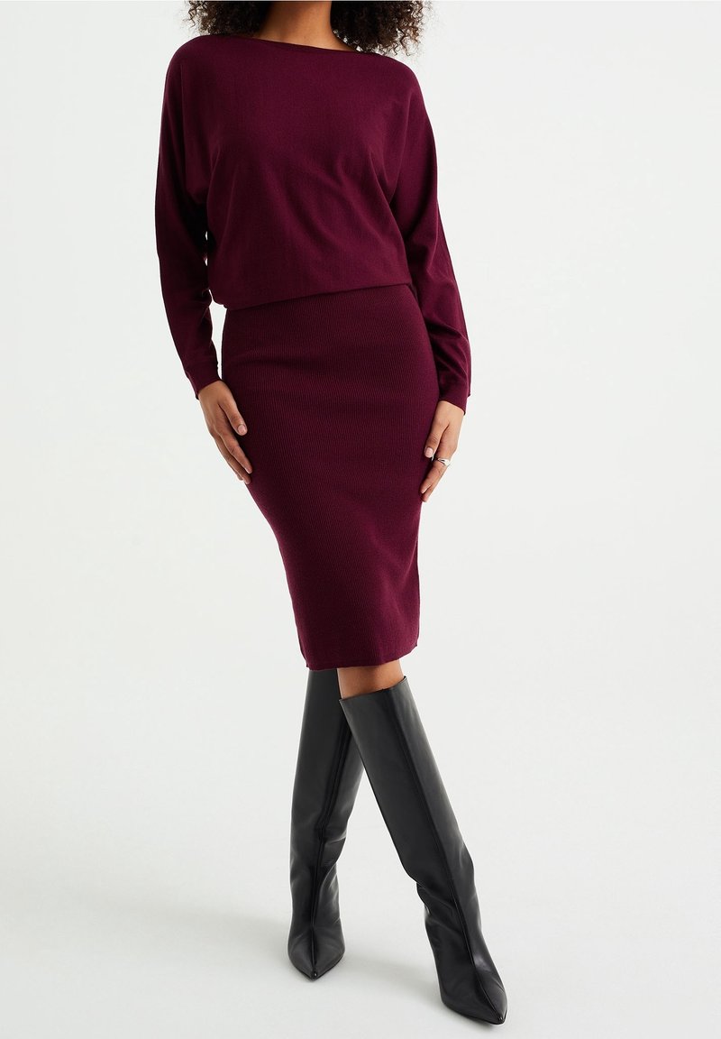 Femme portant une jupe en maille bordeaux ajustée et un haut à manches longues assorti, associée à des bottes noires montantes à pointes, debout les mains sur les hanches.