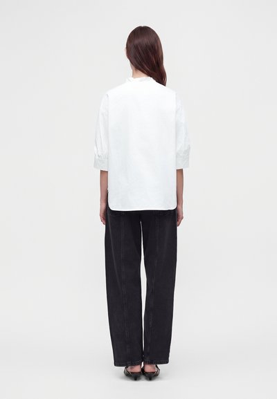 Personne debout de dos portant un chemisier blanc à manches courtes, un pantalon noir à jambes larges et des chaussures noires à talons bas sur un fond uni.