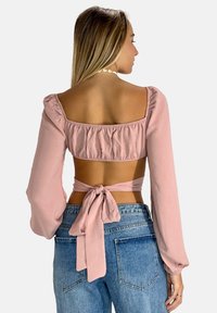 Blusa cropped rosa chiaro con maniche lunghe a palloncino, vita elastica e laccio sul retro. Indossata con jeans in denim blu.