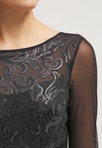 Černé průhledné šaty s vyšívanými, spirálovitými stříbrnými flitry. Klasický kulatý výstřih a dlouhé rukávy zdůrazňují složité detaily a texturu.