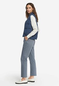 Mörkblå vadderad väst med vertikala sömmar, hög krage, bärs över en ljus färgad långärmad skjorta, ihop med grå byxor och vita loafers.