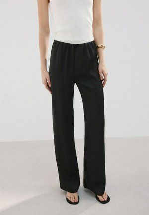 Femme portant un pantalon noir à jambes larges avec une taille élastique, un haut sans manches blanc, des sandales noires et un bracelet en or au poignet droit.