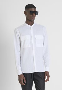 Camicia bianca a maniche lunghe in lino leggero, con due tasche frontali, colletto arrotondato e chiusura con bottoni. Occhiali da sole neri indossati.
