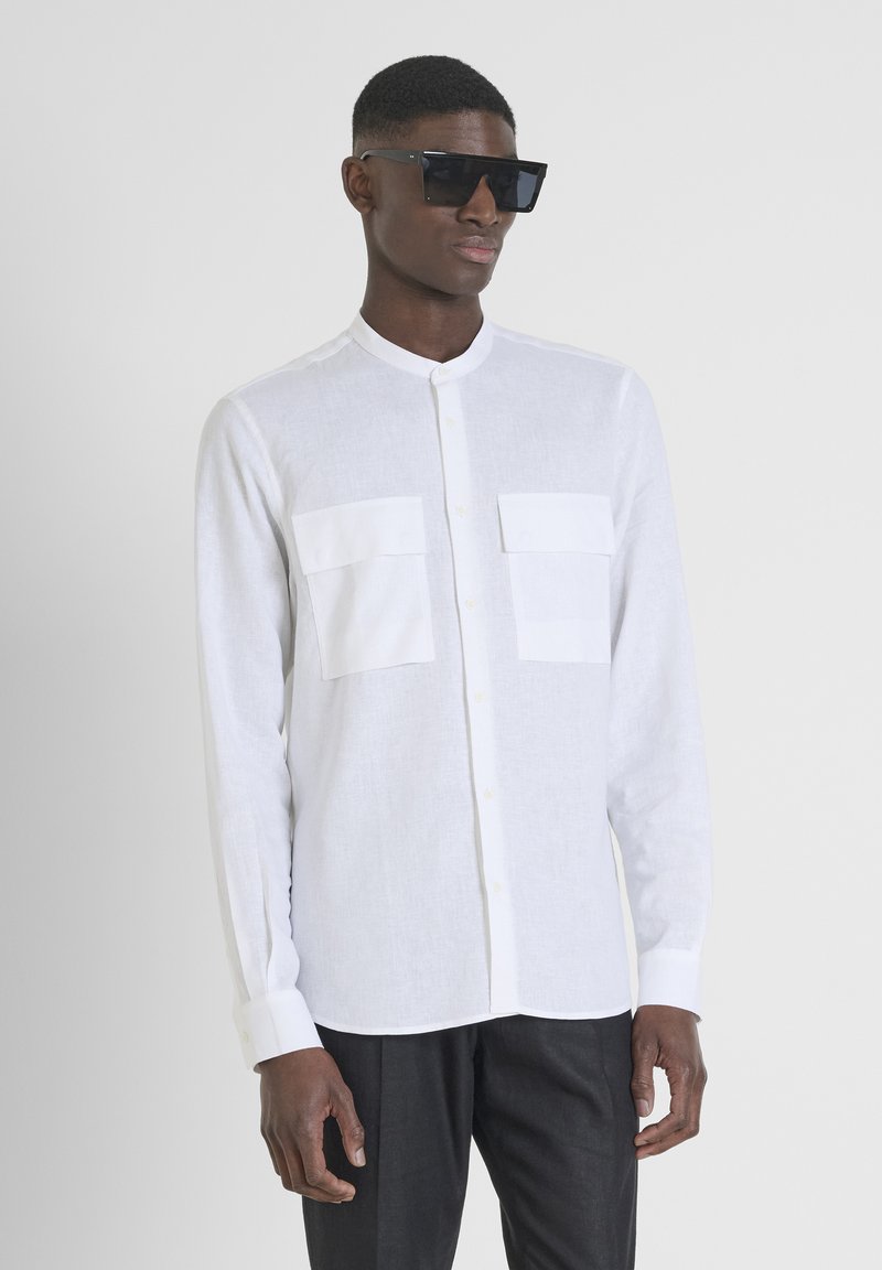 Camicia bianca a maniche lunghe in lino leggero, con due tasche frontali, colletto arrotondato e chiusura con bottoni. Occhiali da sole neri indossati.