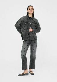 Zwarte spijkerjack en broek met decoratieve zwarte kraal versieringen, ontworpen voor een losse pasvorm. Model draagt patroon platte schoenen.