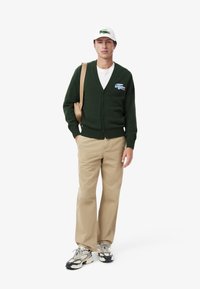 Cárdigan verde con escote en V y botones, que presenta un logotipo bordado. Combinado con pantalones color crema y zapatillas en blanco y negro.