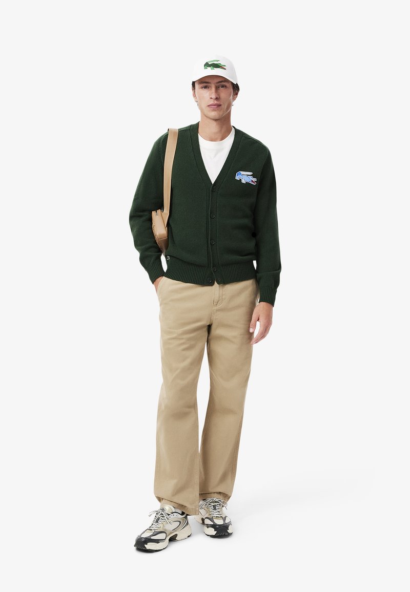 Cárdigan verde con escote en V y botones, que presenta un logotipo bordado. Combinado con pantalones color crema y zapatillas en blanco y negro.
