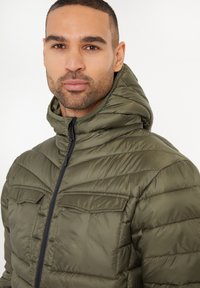 Chaqueta tipo abrigo en verde oliva con capucha, con textura acolchada, dos bolsillos en el pecho y cierre frontal con cremallera.