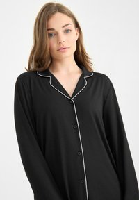 Camisa de pijama negra de manga larga con escote en V, ribete blanco en el collar y botones en la parte delantera, confeccionada en una tela suave.