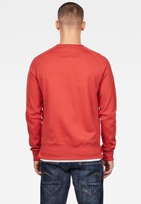 Uomo che indossa una felpa rossa a manica lunga e jeans blu, in piedi con le spalle rivolte verso un semplice sfondo bianco.