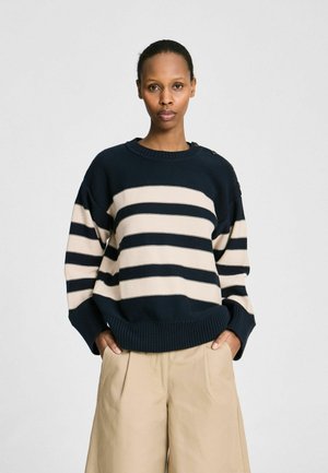 Frau trägt einen navyblauen und beigen gestreiften Pullover mit Knopfdetails und beigen, weit geschnittenen Hosen, steht mit den Händen in den Taschen.