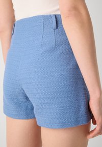 Pantalones cortos azules texturizados con una superficie suave, presentando una cintura de altura media y bolsillos laterales. La tela tiene un patrón sutil.