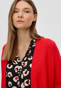 Roter, gestrickter Cardigan über einer schwarzen Bluse mit einem weiß-rot abstrakten Muster. Glatte Textur mit einem lässigen, offenen Design.
