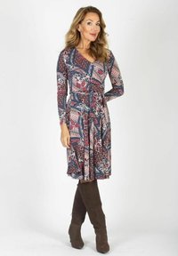 Kobieta stoi z ręką na biodrze, ubrana w długą sukienkę w kwiatowy wzór i ciemne kozaki do kolan, na białym tle.