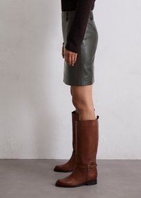 Bottes en cuir marron jusqu'au genou avec une ceinture et un détail de boucle ; associées à une mini-jupe en cuir vert foncé ajustée, montrées de profil.