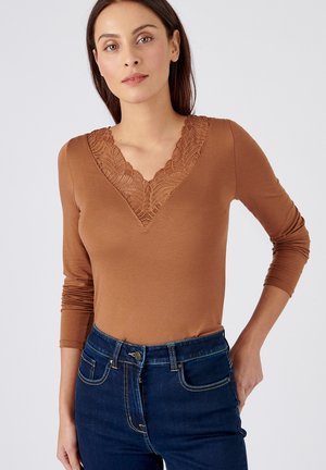 Langsleevige, bruine top met een V-hals en textuurkantdetails, gecombineerd met donkerblauwe jeans met zakken en een knoopsluiting.