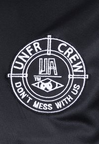 Crna tkanina s okruglim bijelim izvezenim natpisom. Tekst uključuje "UNFR CREW," "UA," i "NE DIRAJTE NAS," s grafikom očiju.