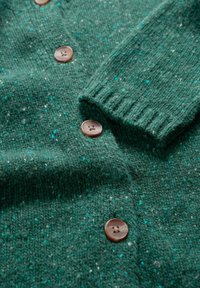 Svetr vyrobený z texturované zelené látky s modrými a bílými skvrnami, vybavený kulatými hnědými knoflíky a ribbenovým detailem na manžetách.
