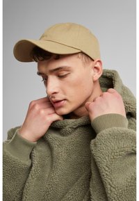 Beige Mütze mit gebogenem Schirm, getragen mit einem strukturierten grünen Fleece-Hoodie. Der Hoodie hat eine lockere Passform und gerippte Bündchen.