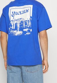 Modrá bavlněná tričko s bílým grafickým designem kaktusů a textem "Dickies" na zadní straně, s krátkými rukávy a uvolněným střihem.