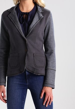 Vrouw draagt een donkergrijze getextureerde blazer over een marineblauwe blouse met striksluiting, gecombineerd met donkerblauwe aansluitende jeans.