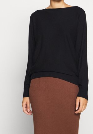 Femme portant un haut à manches longues noir et une jupe côtelée marron taille haute, debout devant un fond blanc uni.