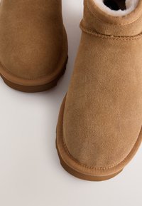 Paire de bottes d'hiver en suède beige avec coutures visibles et doublure en polaire blanche sur un fond clair, vues de dessus.