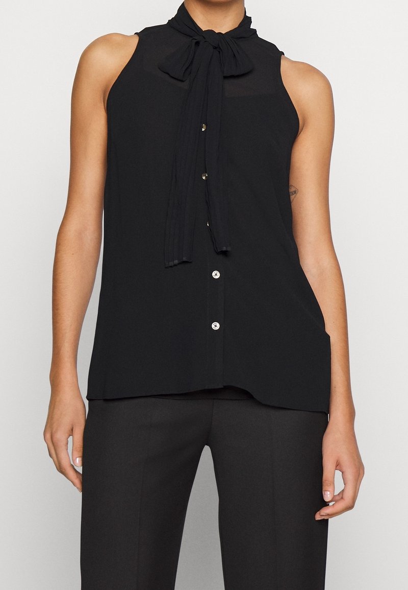 Blouse noire sans manches avec boutons sur le devant et foulard noué autour du cou, portée avec un pantalon noir.