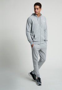 Grå zip-up hoodie och matchande sweatpants tillverkade av mjukt material. Huvtröja och framfickor detaljerade; ihopparat med svarta och grå sneakers.