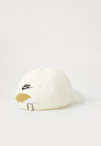 Balta medvilnės kepurė su kreivu kraštu, priekyje puikuojasi juodas Nike swoosh logotipas, o gale yra reguliuojama metalinė juostelė.