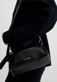 Borsa a mano in pelle nera con una texture liscia, chiusura con zip e hardware argentato. Presenta un sottile logo Calvin Klein sul davanti.