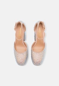 Des talons de sandales en matériau nude ornés de strass irisés, avec un design à bout rond et une sangle autour de la cheville pour le soutien.