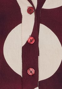 Maroon och krämfärgad mönstrad skjorta med knappar framtill, som har stora cirkulära mönster, med röda knappar och ett strukturerat, vävt tyg.