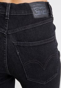 Jeans de mezclilla negra con una cintura alta. Presenta un parche de cuero en la parte posterior con la marca, dos bolsillos traseros y detalles de costura sutiles.