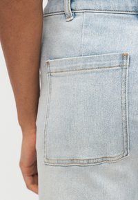 Κοντινή λήψη πίσω τσέπης από γαλάζιο τζιν με ορατή ραφή και μερικώς ορατό χέρι με σκούρο τόνο δέρματος δίπλα του.