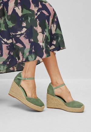 Jambes portant des espadrilles compensées en daim vert avec des brides à la cheville et une jupe colorée à motif abstrait.